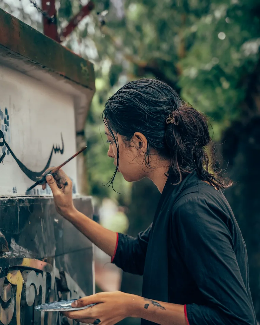 Persona pintando un trazo caligráfico con pincel fino sobre un panel claro, sosteniendo una paleta con la otra mano