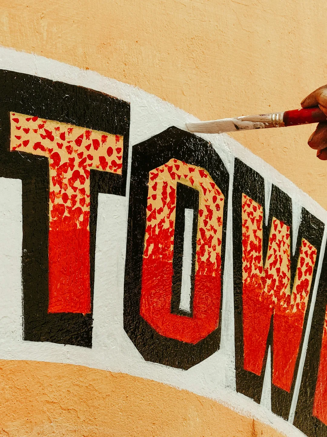 Pincel de rotulista repasando el perfil blanco de unas letras rojas y negras pintadas sobre un muro enfoscado